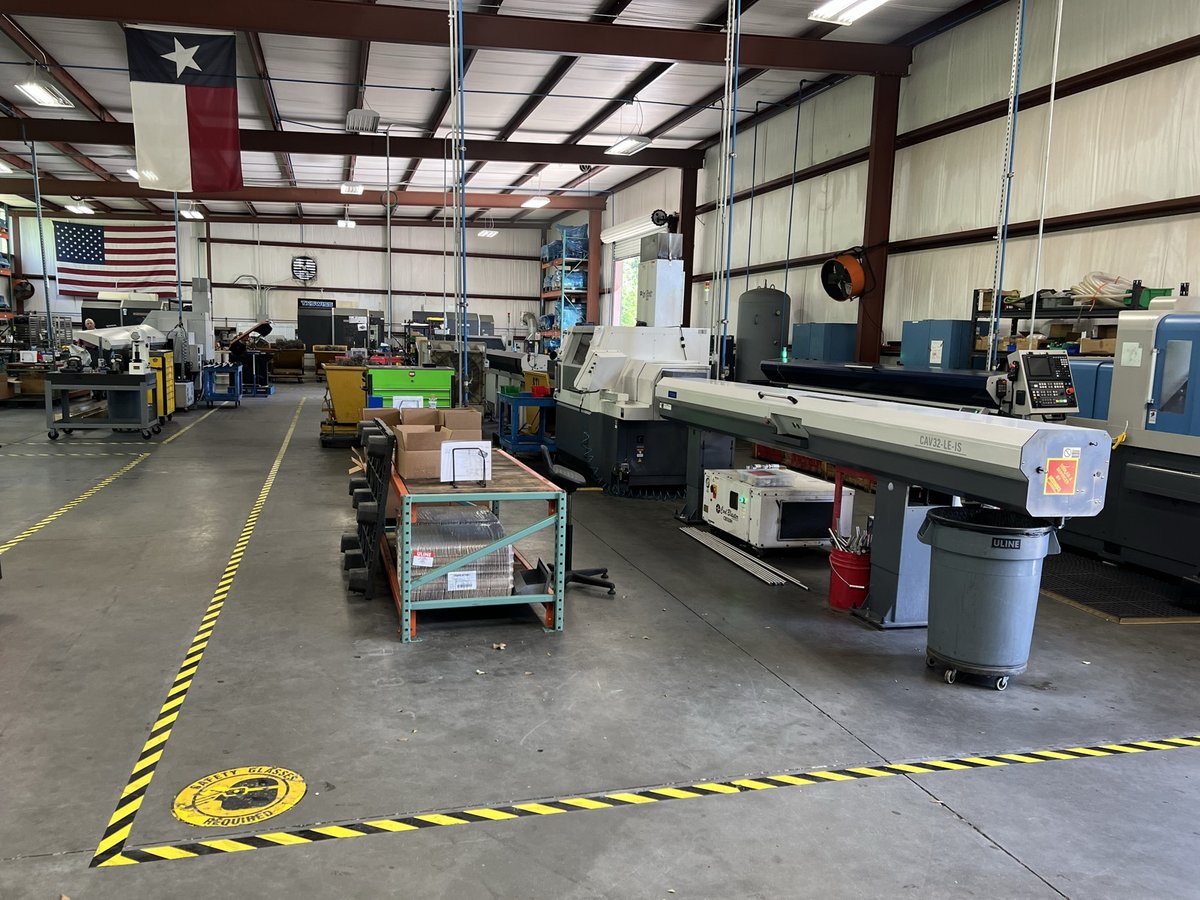 TXSwiss precision Swiss CNC shop floor in Conroe, Texas with bar-fed lathes, racks of bar stock, and US and Texas flags overhead — ISO 9001:2015 certified manufacturing facility